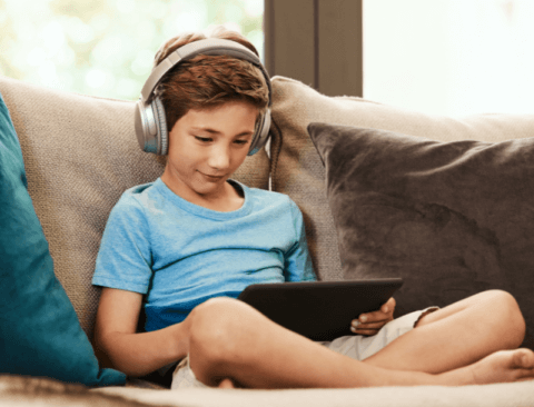 Ein Junge sitzt auf dem Sofa, hat Kopfhörer an und schaut auf sein Tablet.