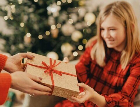 Ein Mädchen in einem rot karierten Kleid lächelt und erhält ein Geschenk in goldenem Papier mit rotem Band von einer Person in einem roten Pullover. Im Hintergrund steht ein geschmückter Weihnachtsbaum mit Lichtern und Ornamenten.