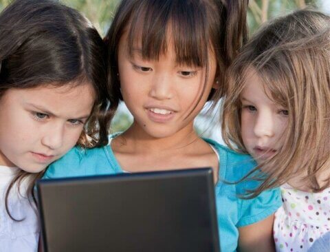 Drei Kinder schauen gemeinsam auf einen Laptop-Bildschirm. Sie wirken interessiert und konzentriert. Die Szene scheint bei Tageslicht im Freien aufgenommen worden zu sein.