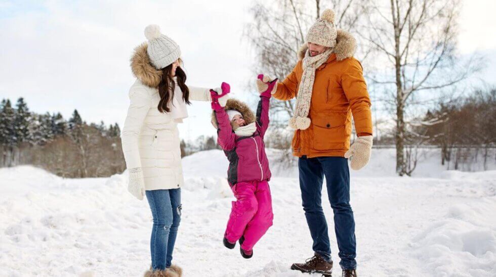 Eine Familie spielt im Schnee. Ein Mann in einer orangefarbenen Jacke und eine Frau in einer weißen Jacke halten ein fröhliches Kind, das in einem pinken Schneeanzug in die Luft springt. Der Hintergrund zeigt verschneite Bäume und blauen Himmel.